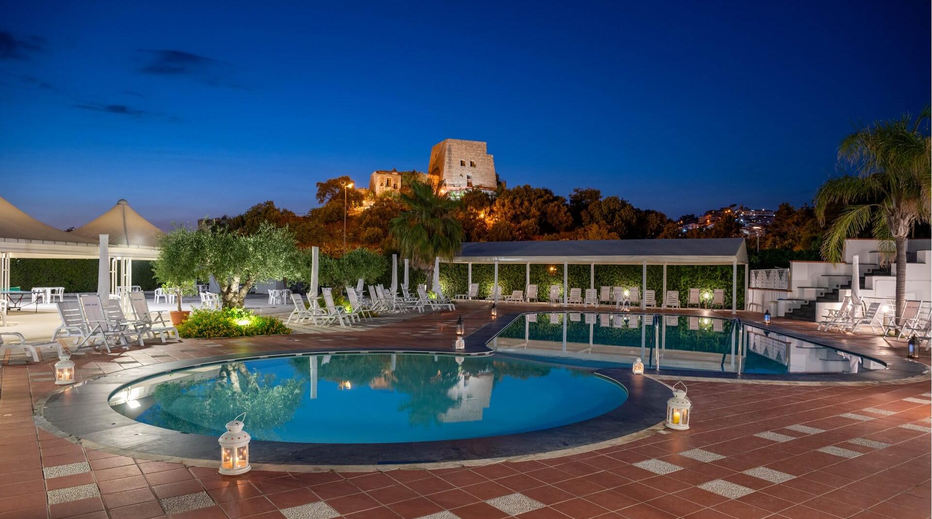 Piscina illuminata di sera con vista su antiche rovine su una collina.