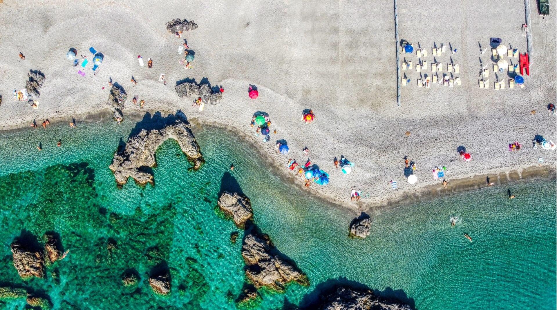 Spiaggia con ombrelloni colorati, mare cristallino e rocce, vista dall'alto.