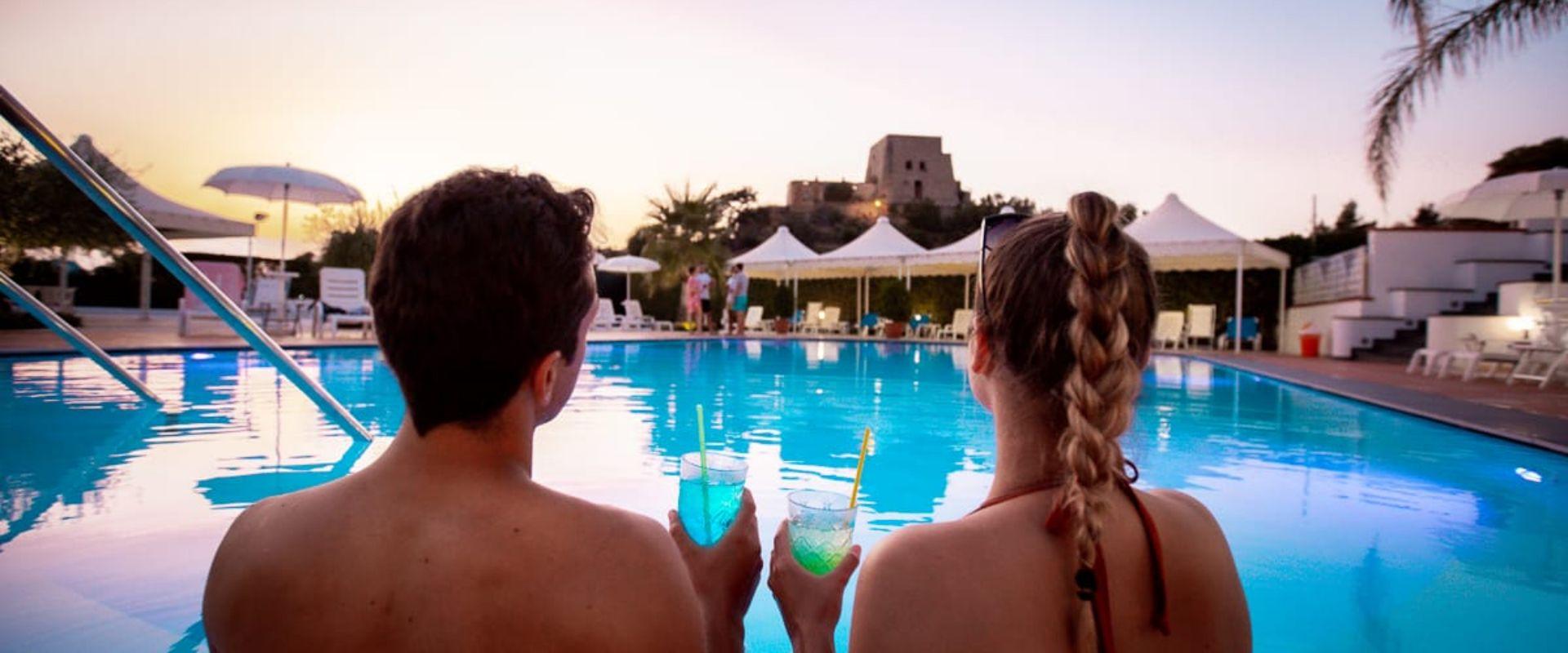 Coppia rilassata in piscina al tramonto con cocktail in mano.