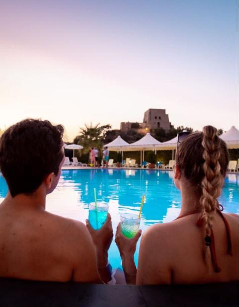 Coppia rilassata in piscina con drink al tramonto, sfondo di un castello.