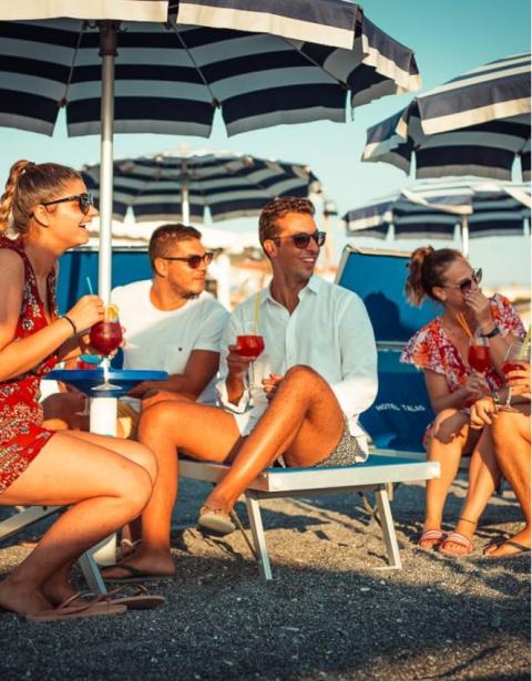 Gruppo di amici in spiaggia, sotto ombrelloni, con drink in mano, sorridono al sole.