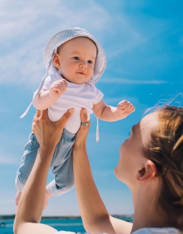 Bambino con cappello sollevato da una donna sotto un cielo azzurro.