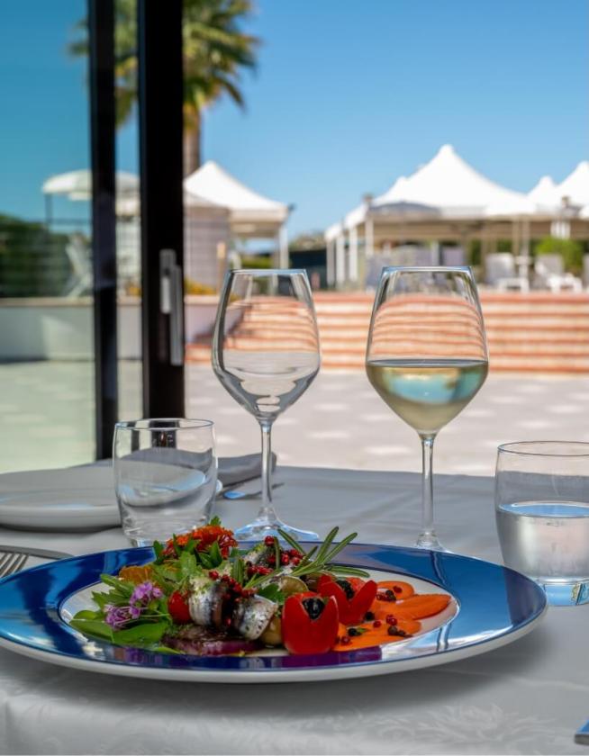 Piatto elegante con pesce e verdure, bicchieri di vino, vista piscina e tende bianche.